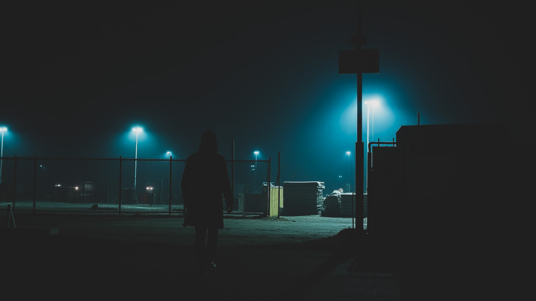 A shadowy figure sneaking near a fenced construction site at night, partially illuminated by security lights, emphasizing material theft prevention in construction.